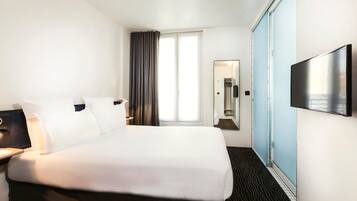 Habitación de hotel con una cama grande, un televisor de pantalla plana montado en la pared, una ventana con cortinas, un espejo y una mesita de noche con lámpara.