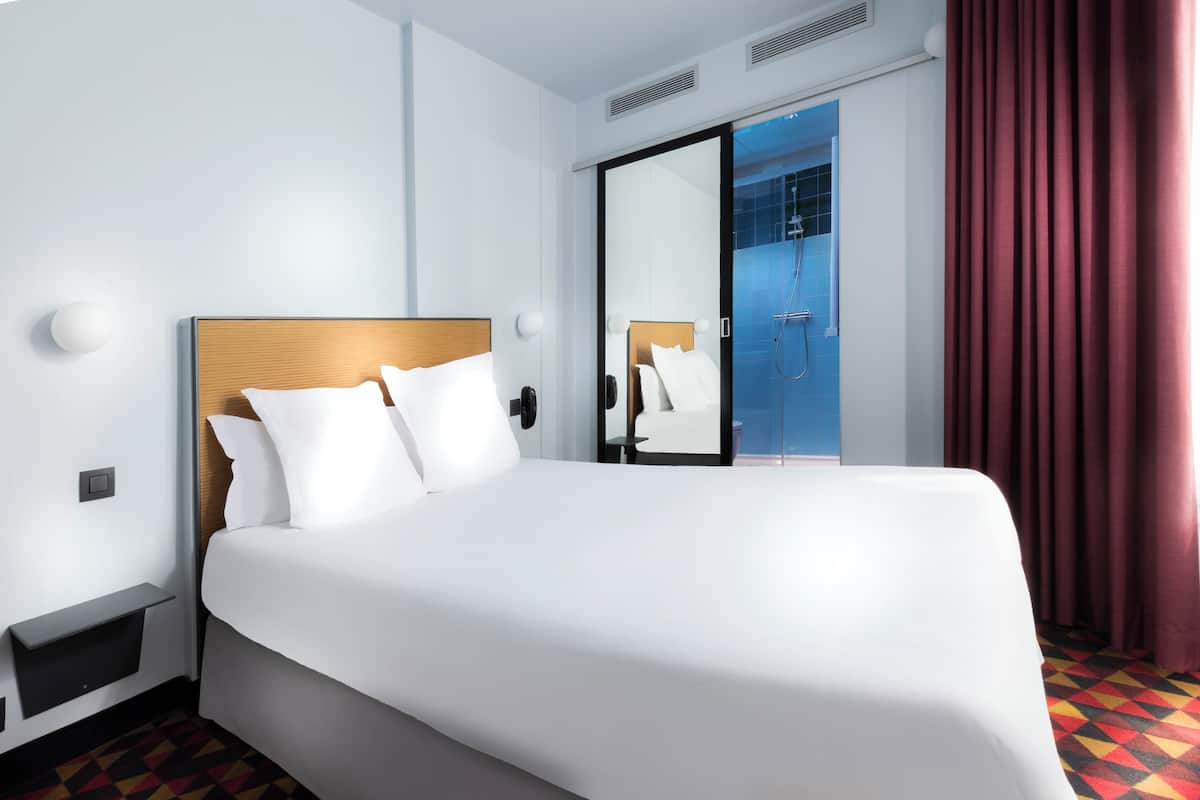 A hotel room with a large bed, a bedside table, a mirror, a shower area, and a red curtain.