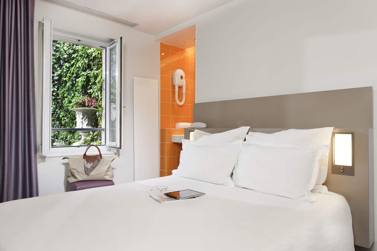 A hotel room with a bed, a window with curtains, a bedside table with a handbag, and a wall-mounted phone.