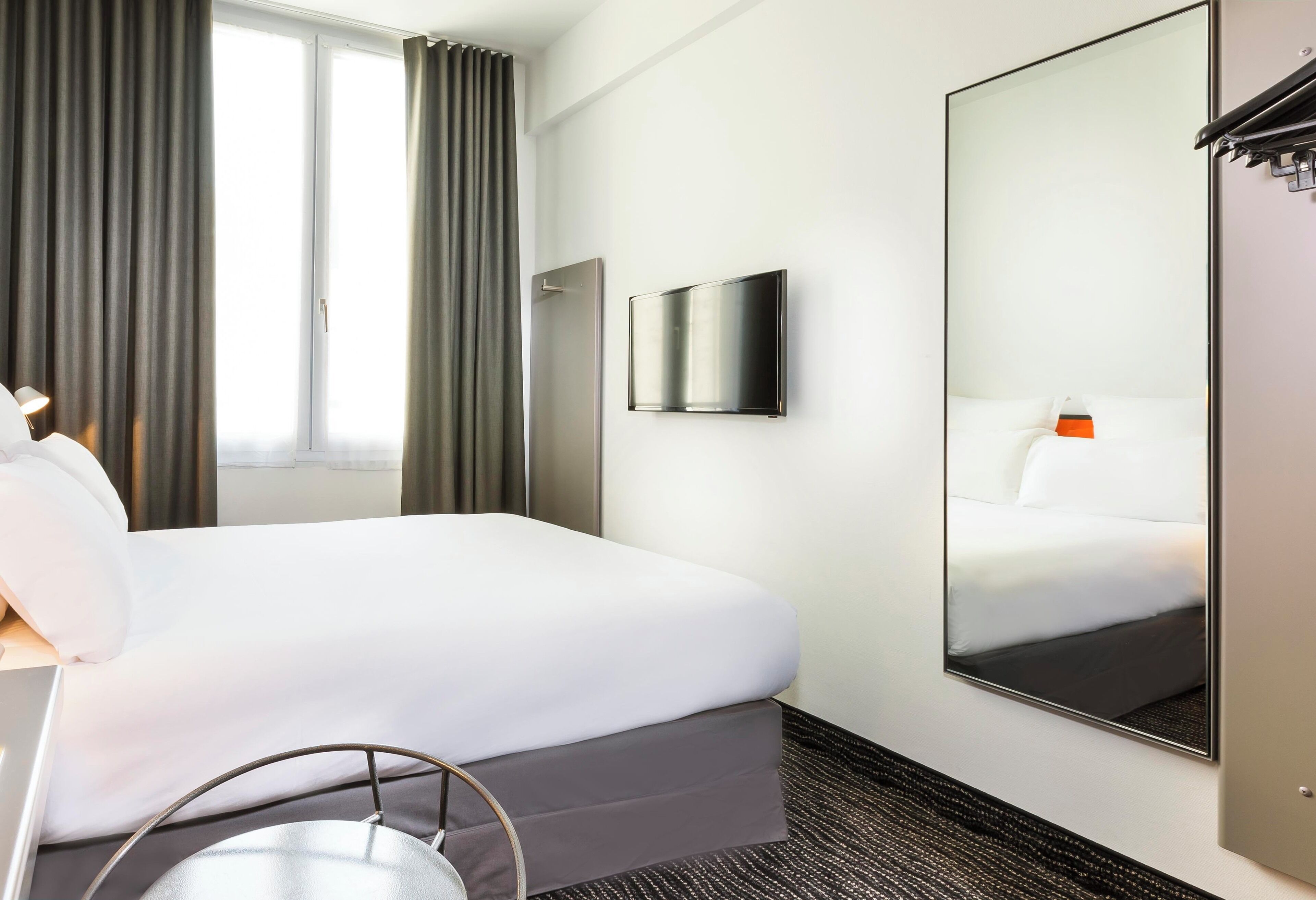 Quarto de hotel com uma cama, televisão de tela plana, espelho grande e janela com cortinas.