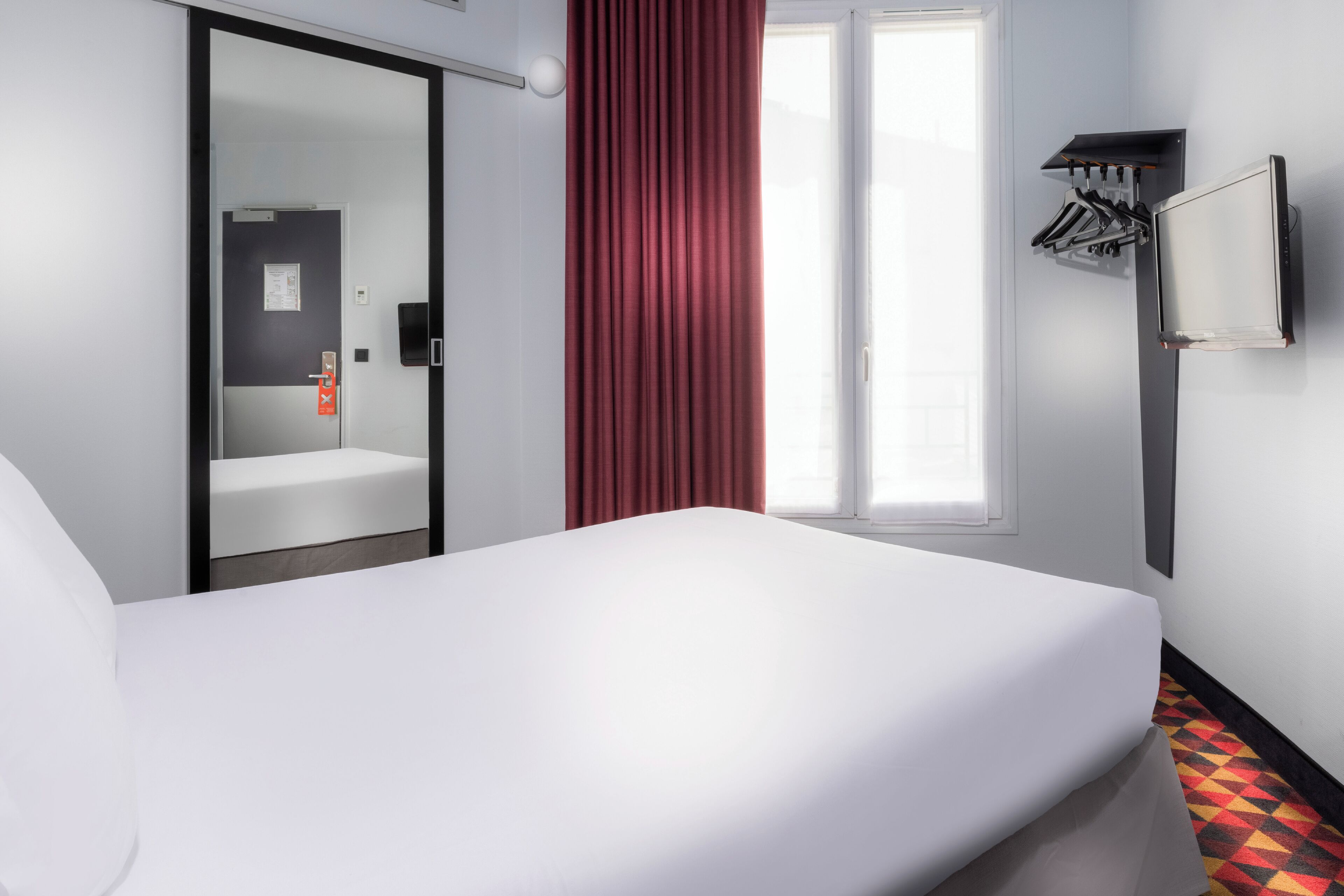 A hotel room with a bed, a mirror, a red curtain, a television, and a coat rack.