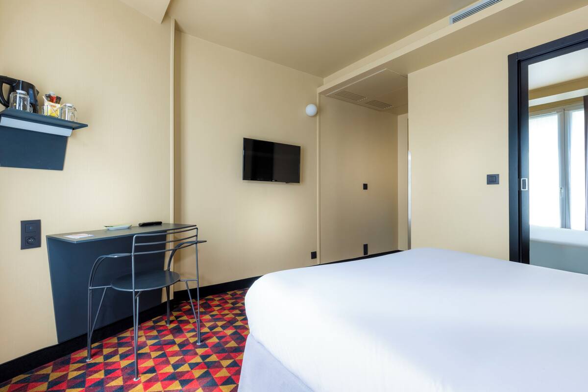 A hotel room with a bed, a desk with a chair, a television on the wall, and a colorful patterned floor.