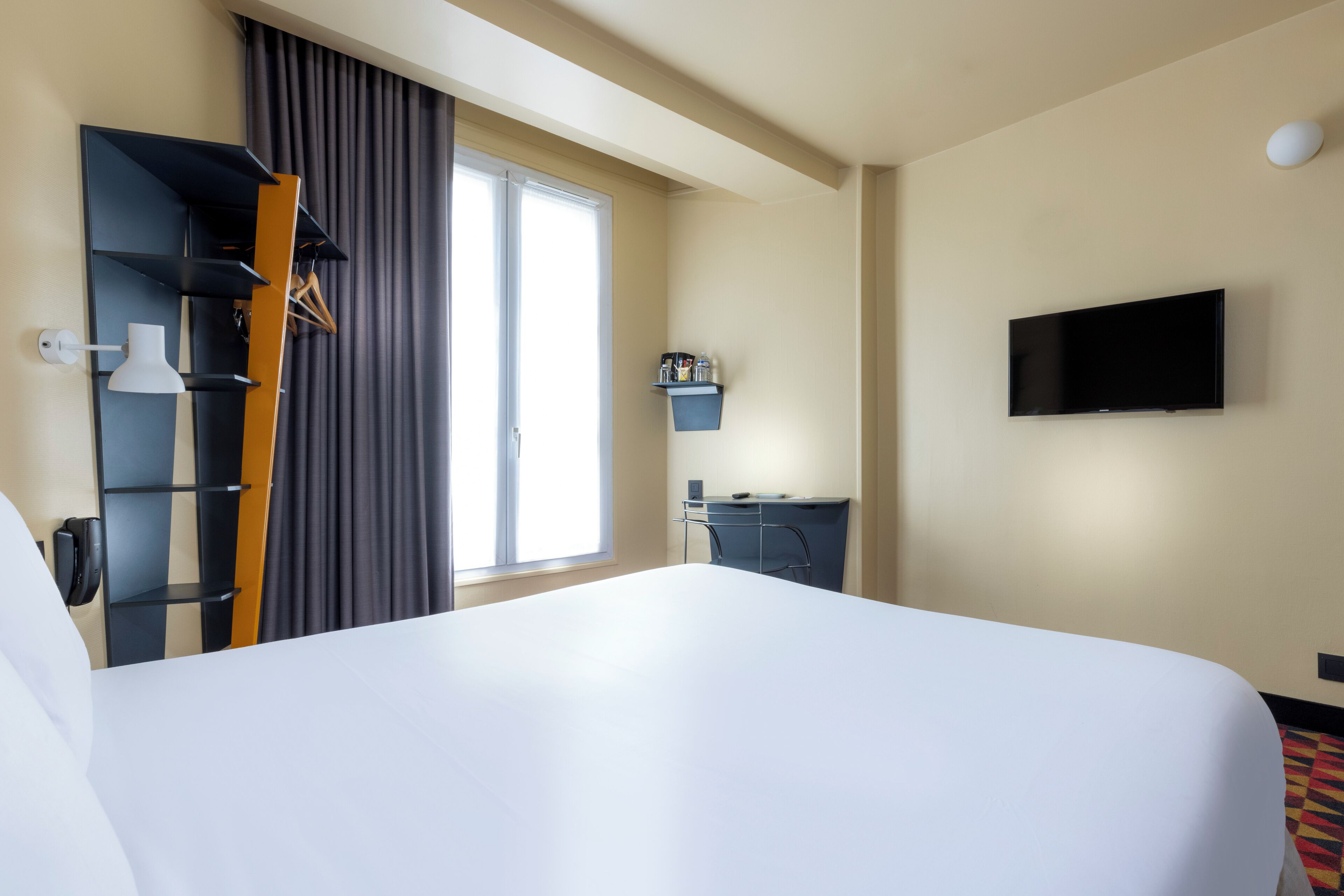 Habitación de hotel con una cama grande, televisor de pantalla plana montado en la pared, escritorio con silla, perchero y ventana con cortinas.