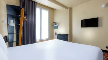 Habitación de hotel con una cama grande, televisor de pantalla plana montado en la pared, escritorio con silla, perchero y ventana con cortinas.
