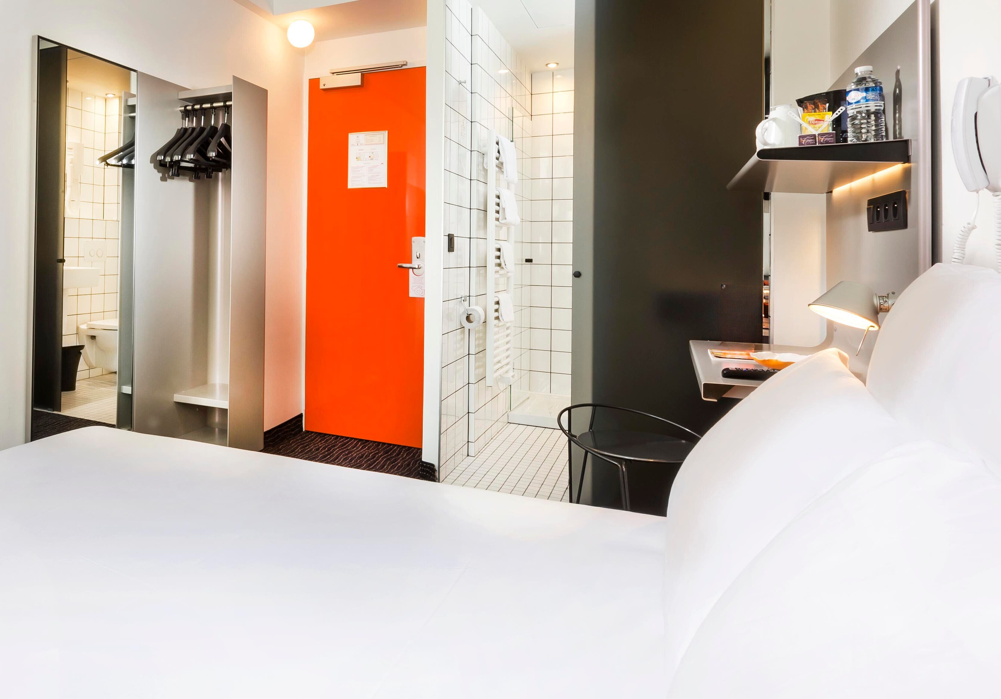 A hotel room with a white bed, an orange door, a shower area, and a small table with a lamp.