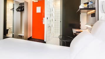 Une chambre d’hôtel avec un lit blanc, une porte orange, un espace douche et une petite table avec une lampe.