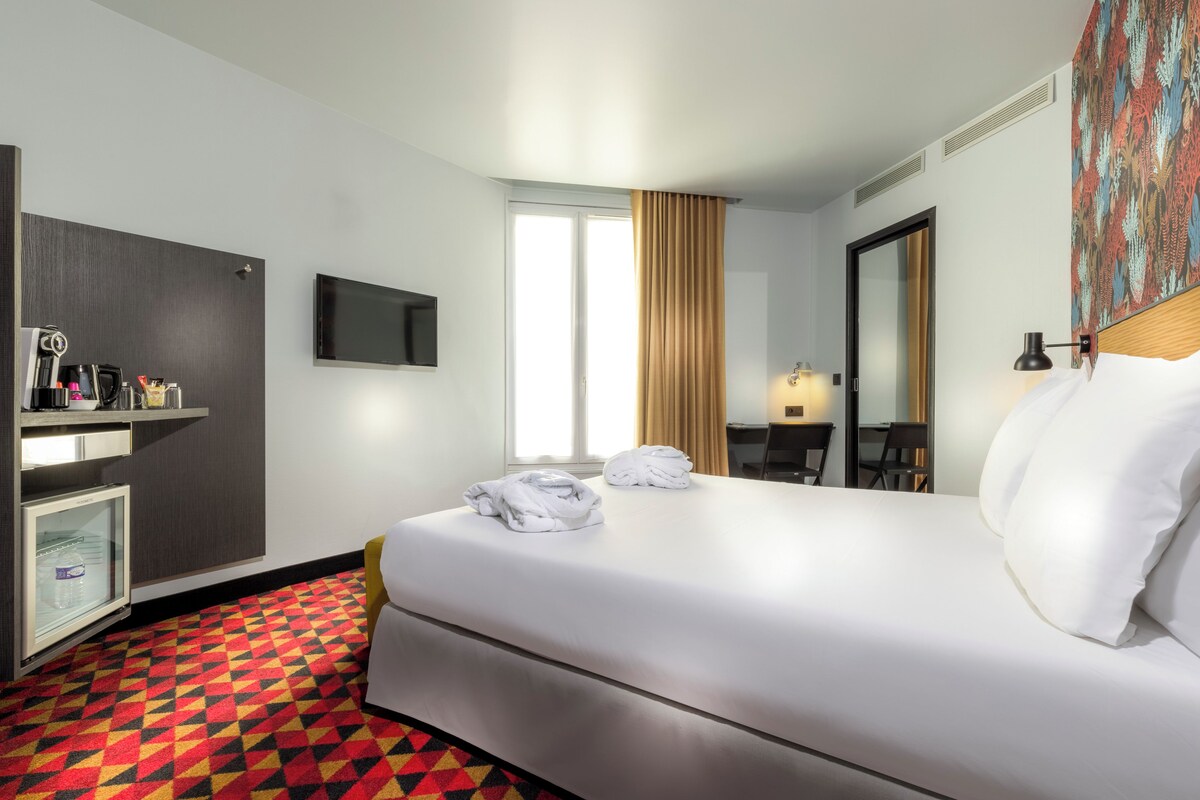 A hotel room with a bed, a television, a desk with a chair, a mirror, and a patterned carpet.