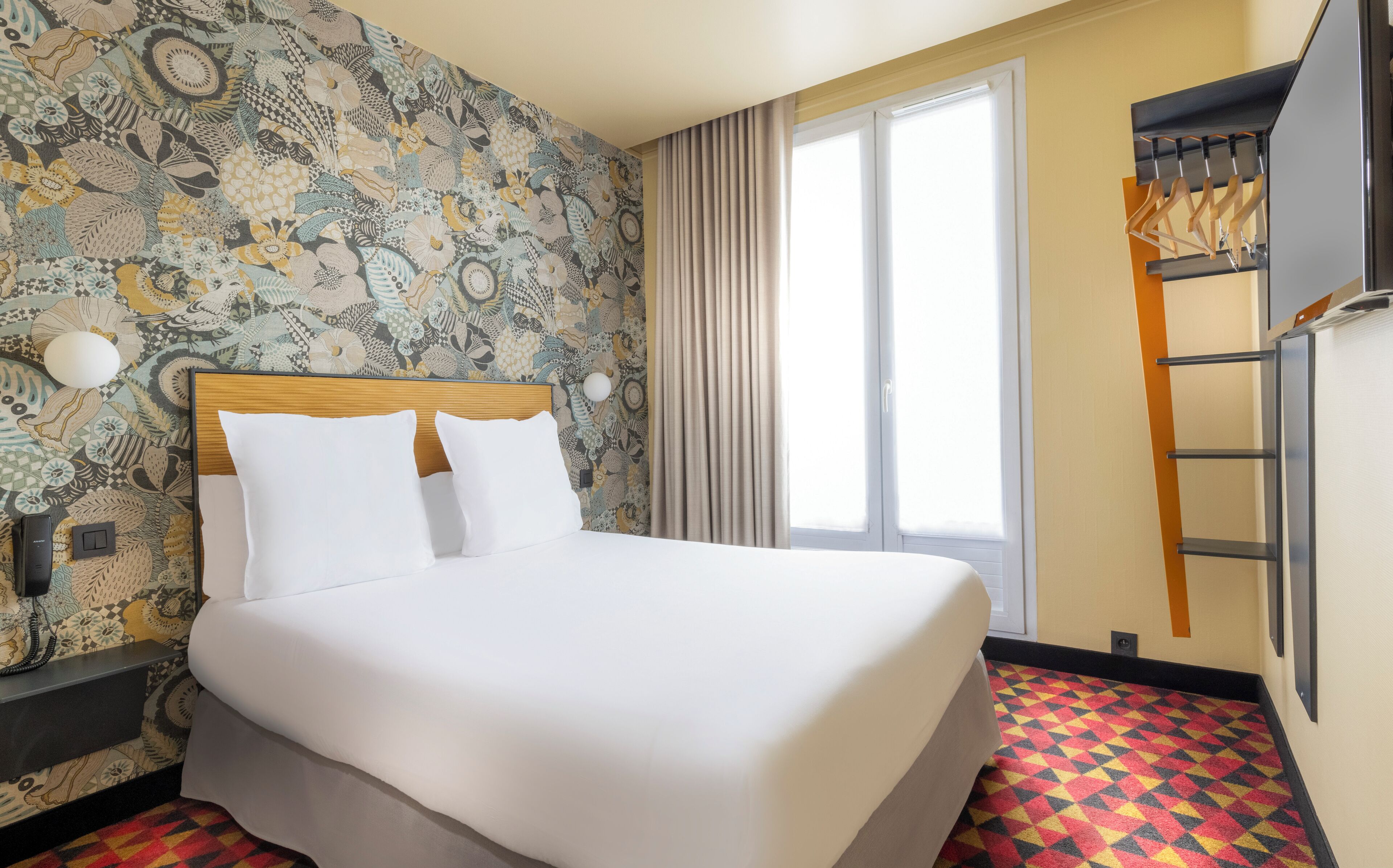 Une chambre d’hôtel avec un papier peint à motifs, un lit avec du linge de maison blanc, une fenêtre avec des rideaux, une échelle en bois et un tapis à motifs rouges et jaunes.