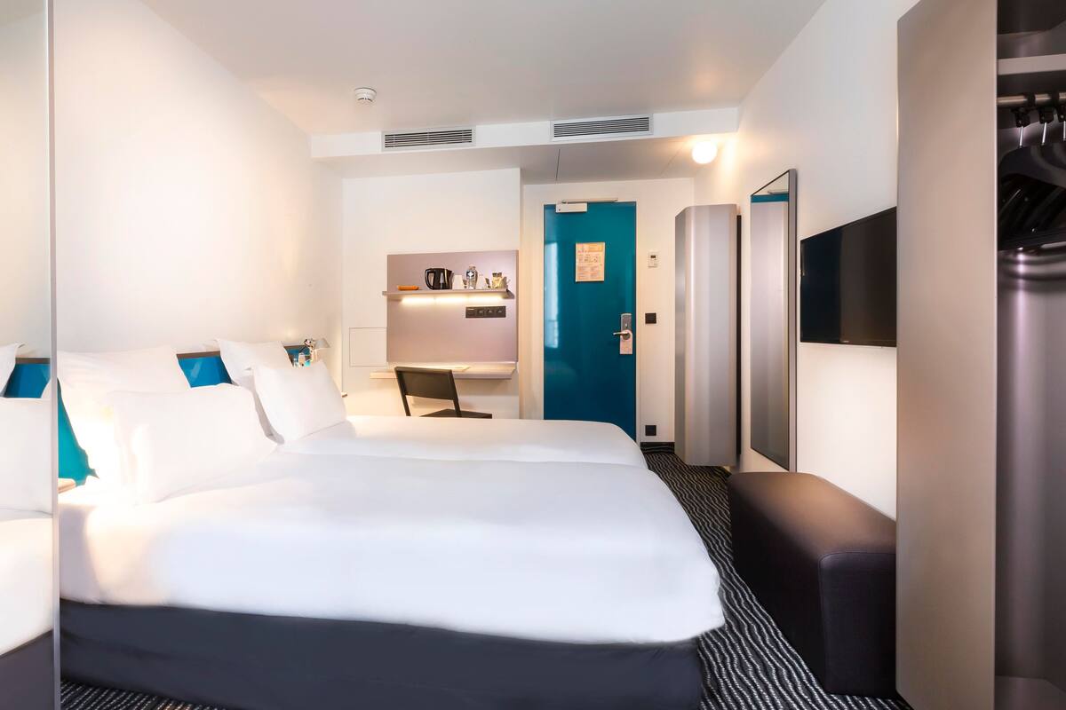 A hotel room with a large bed, a desk, a chair, a wardrobe, a mirror, and a door.