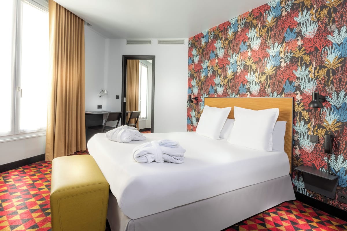 A hotel room with a large bed, a yellow ottoman, a desk, and a vibrant floral wallpaper.