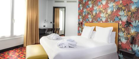 A hotel room with a large bed, a yellow ottoman, a desk, and a vibrant floral wallpaper.
