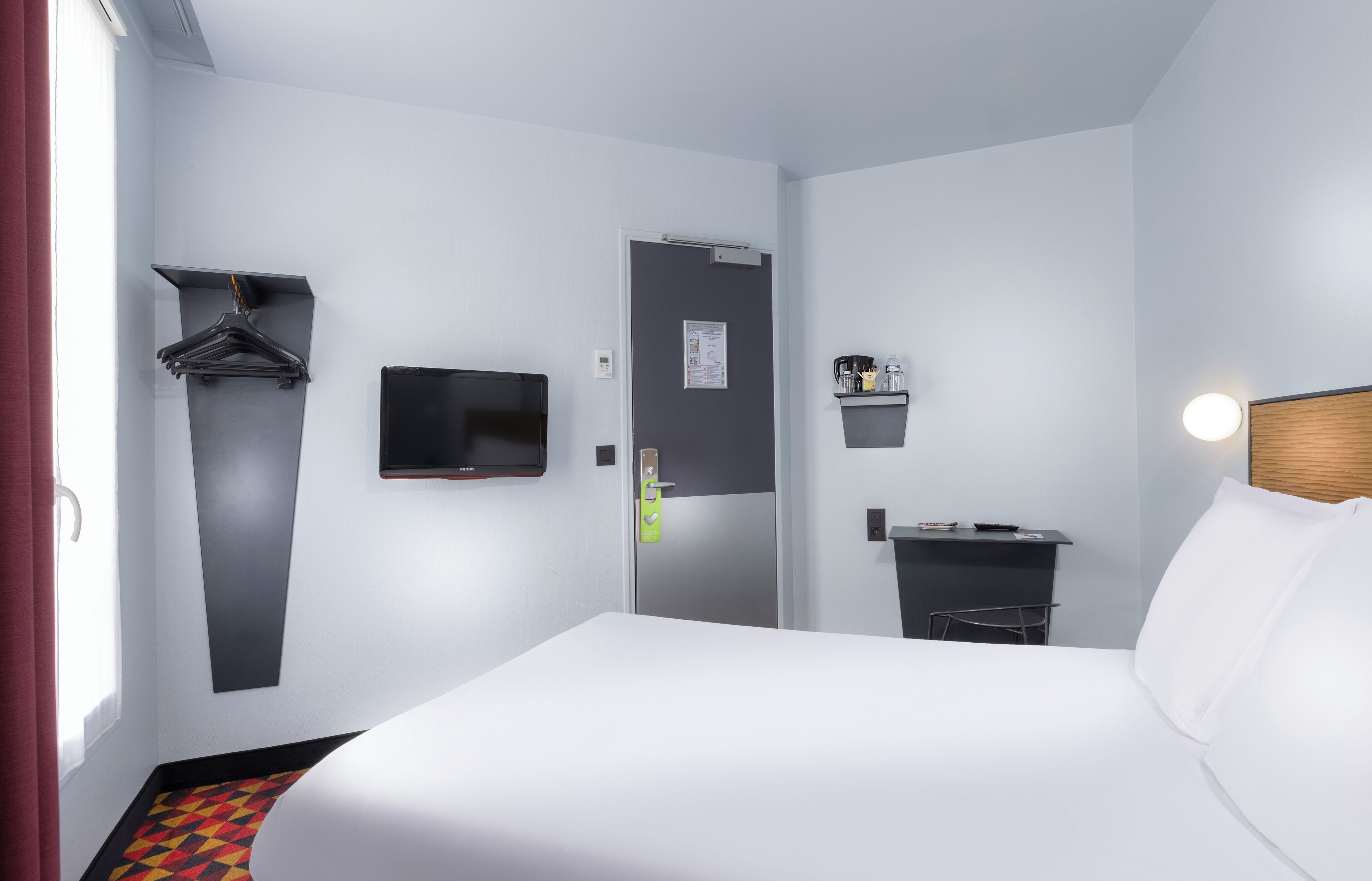 Habitación de hotel con una cama, televisor, escritorio y silla, perchero y un estante pequeño con artículos de aseo.