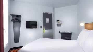 Habitación de hotel con una cama, televisor, escritorio y silla, perchero y un estante pequeño con artículos de aseo.
