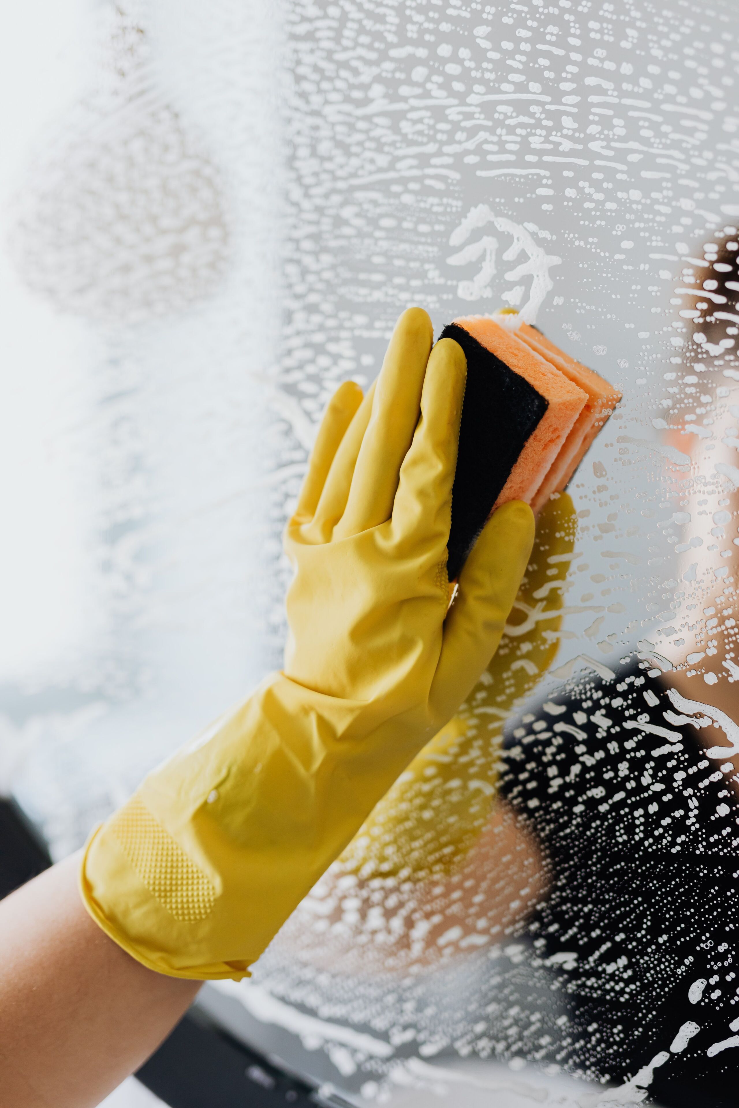 A hand wearing a yellow rubber glove holding a cleaning sponge against a glass surface with water droplets.