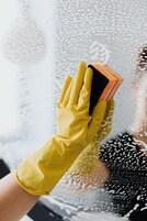 Una mano con un guante de goma amarillo sostiene una esponja de limpieza contra una superficie de vidrio con gotas de agua.