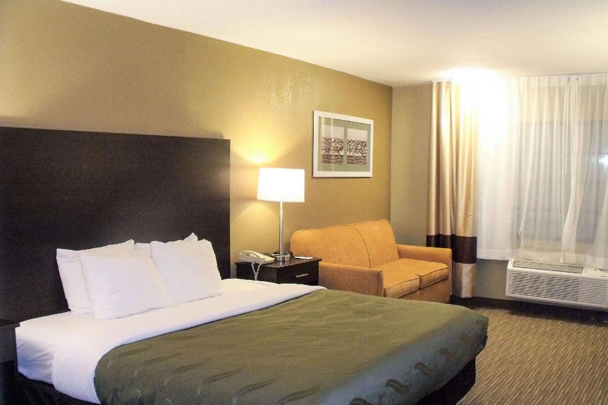 Une chambre d’hôtel avec un lit, une chaise, une lampe, un téléphone et un tableau au mur.