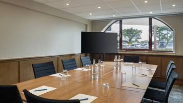 Uma sala de conferências com uma mesa comprida, cadeiras, uma tela grande e vista para um estacionamento pelas janelas.