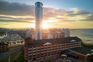 Un edificio de gran altura con una torre cilíndrica, rodeado de otros edificios y un frente marítimo.