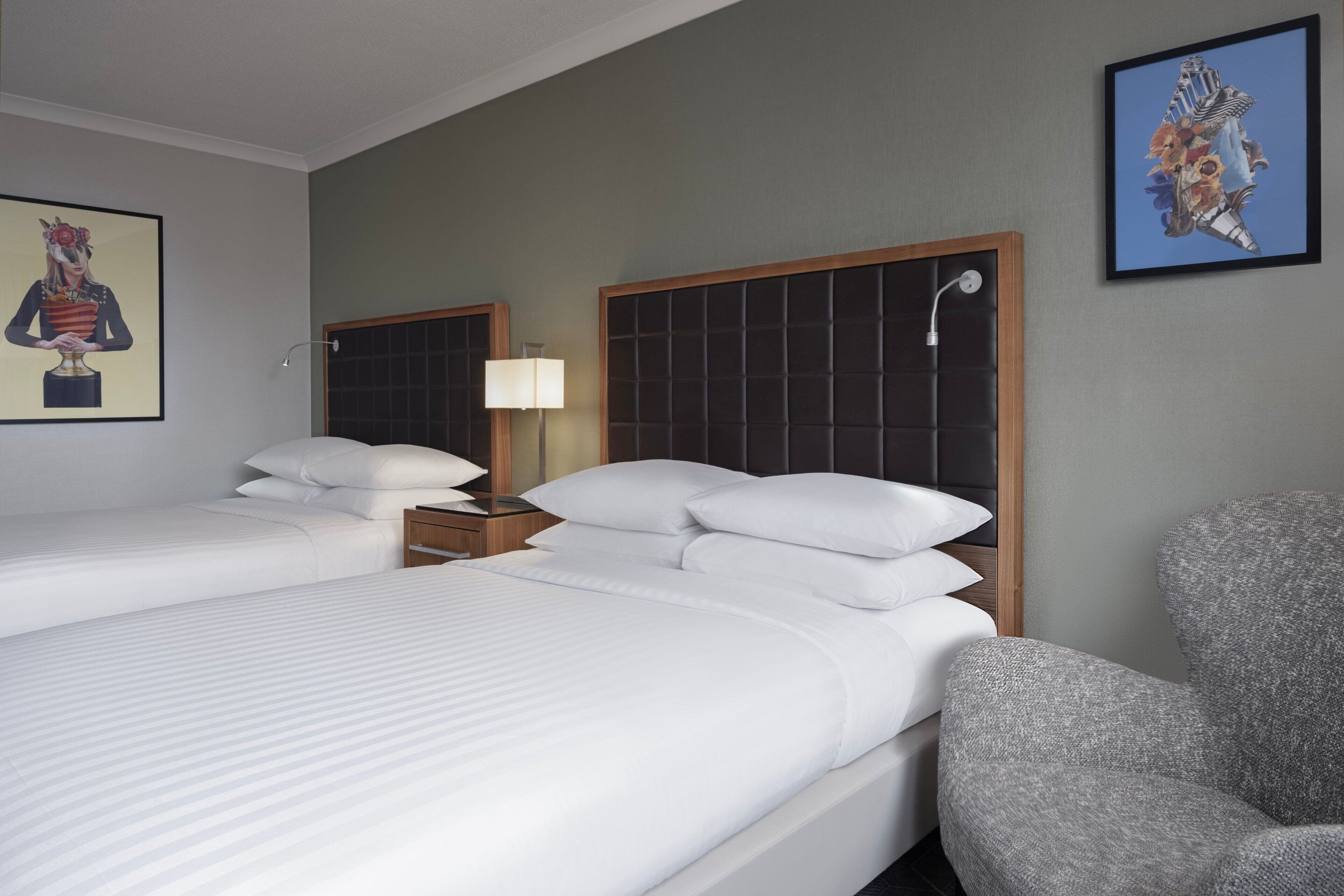 Habitación de hotel con dos camas, una mesita de noche, una silla y cuadros en las paredes.