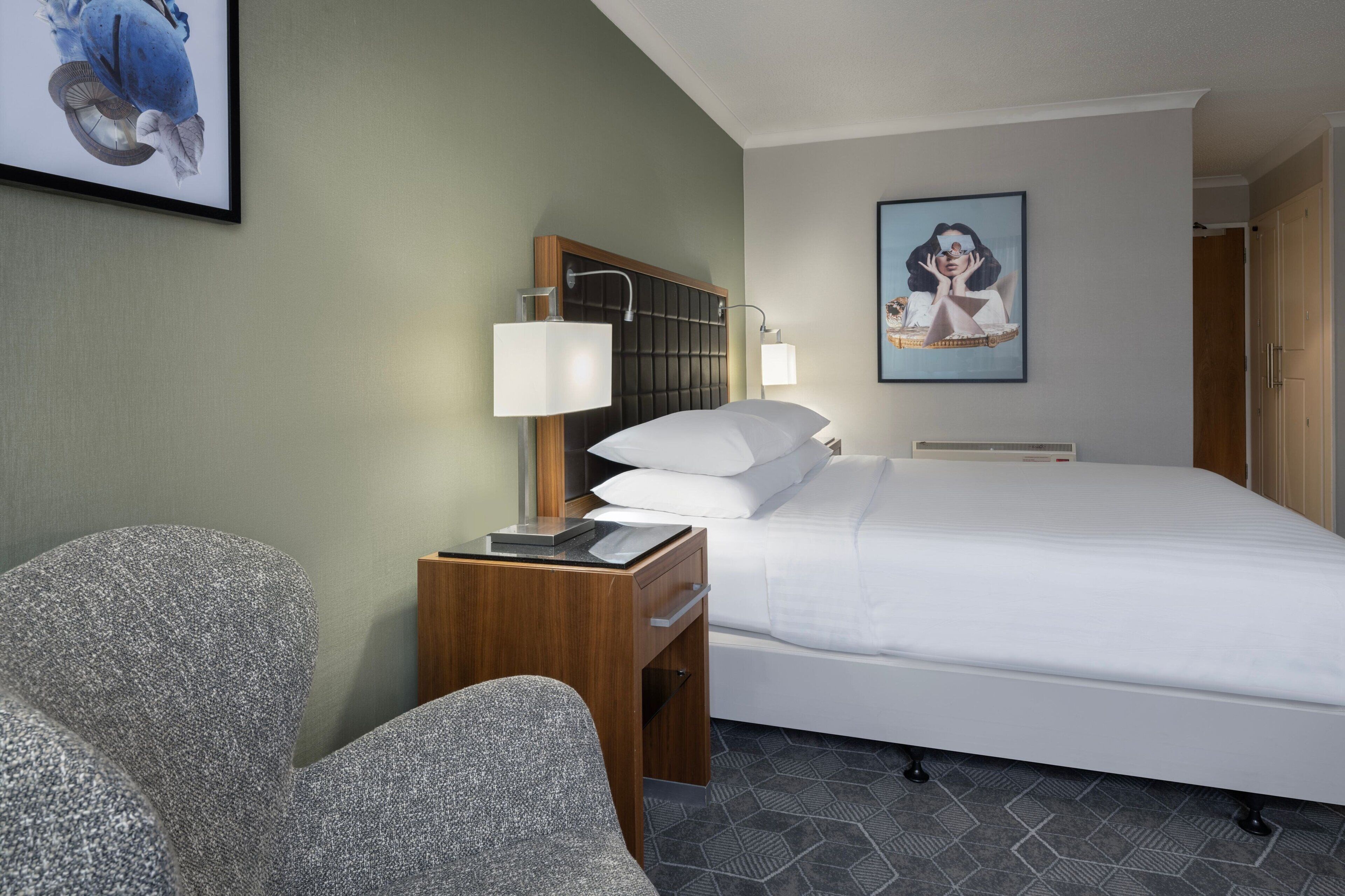Ein Hotelzimmer mit einem Bett, einem Schreibtisch mit Lampe, einem Sessel und einem Bild an der Wand.