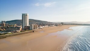 Un paisaje costero con una playa extensa de arena, un edificio moderno alto y un fondo montañoso.