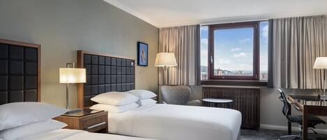 A hotel room with two beds, a desk, a chair, and a view of the city.