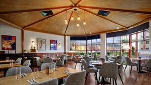 Un restaurante espacioso con techos altos, vigas de madera y amplios ventanales que ofrecen vistas del paisaje urbano. Las mesas están ordenadas con sillas modernas.