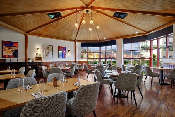 A spacious restaurant with high ceilings, wooden beams, and large windows offering views of the cityscape. Tables are neatly arranged with modern chairs.