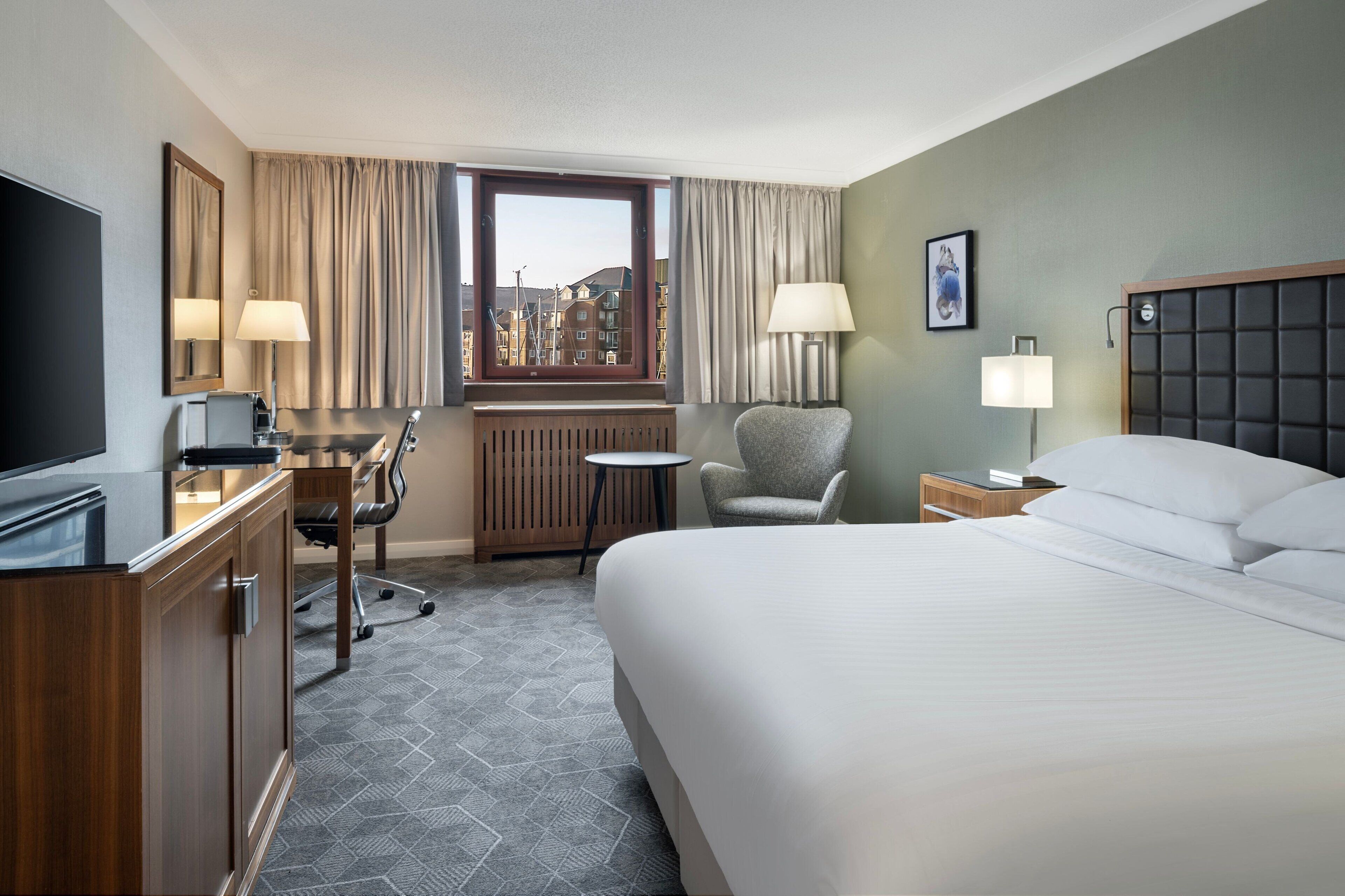 A hotel room with a large bed, a desk with a computer, a chair, a small table, and a window with curtains.