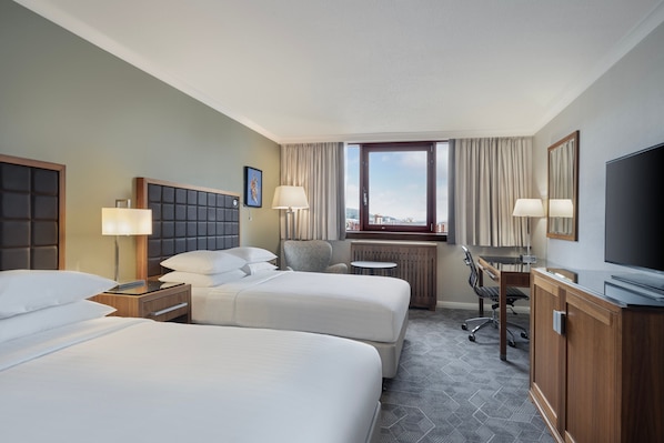 A hotel room with two beds, a desk, a chair, a TV, and a window with curtains.