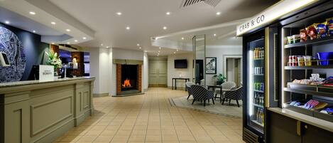 A hotel lobby with a reception desk, a fireplace, a dining area, and a grab-and-go food station.
