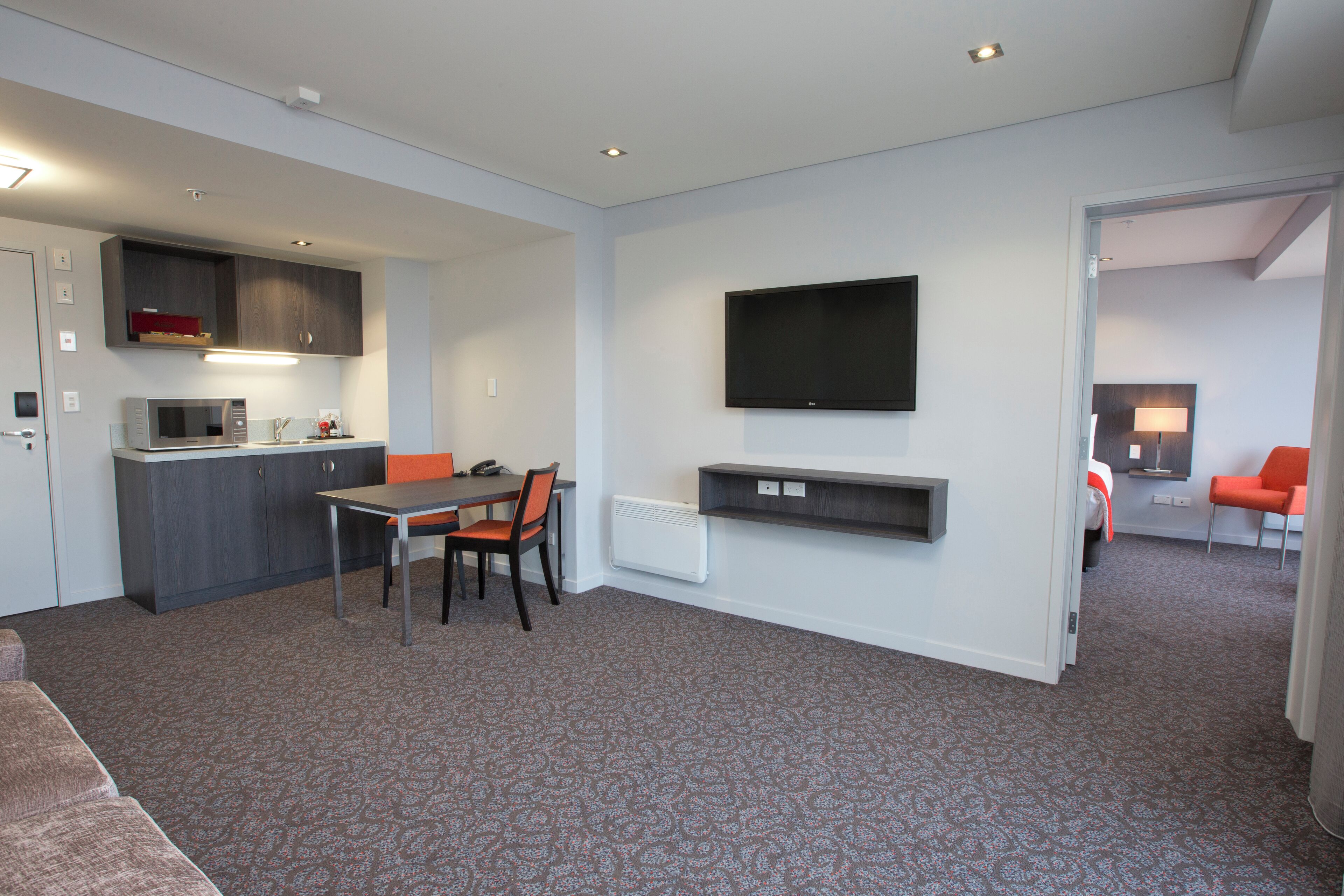 A modern hotel room with a kitchenette, a dining area, a TV mounted on the wall, and a seating area with a sofa and chairs.