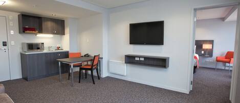 A modern hotel room with a kitchenette, a dining area, a TV mounted on the wall, and a seating area with a sofa and chairs.