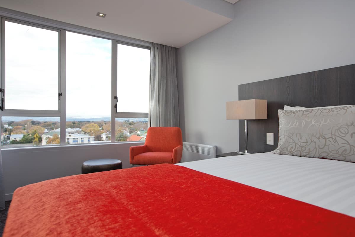 Ein Hotelzimmer mit einem großen Bett, einem Sessel, einer Lampe und Blick auf die Stadt durch das Fenster.