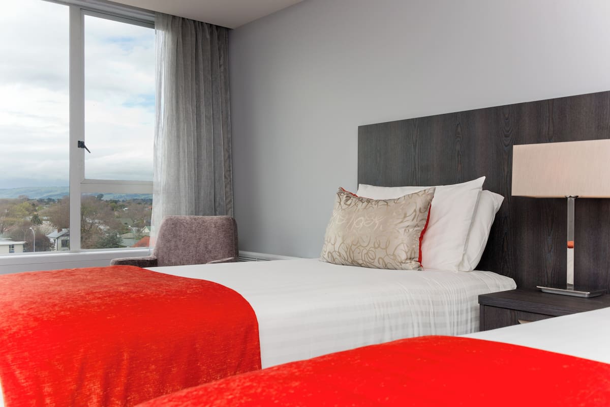 Ein Hotelzimmer mit großem Fenster, einem Bett mit weißer Bettwäsche, einem roten Akzent, einem Sessel, einem Nachttisch und einer Lampe.