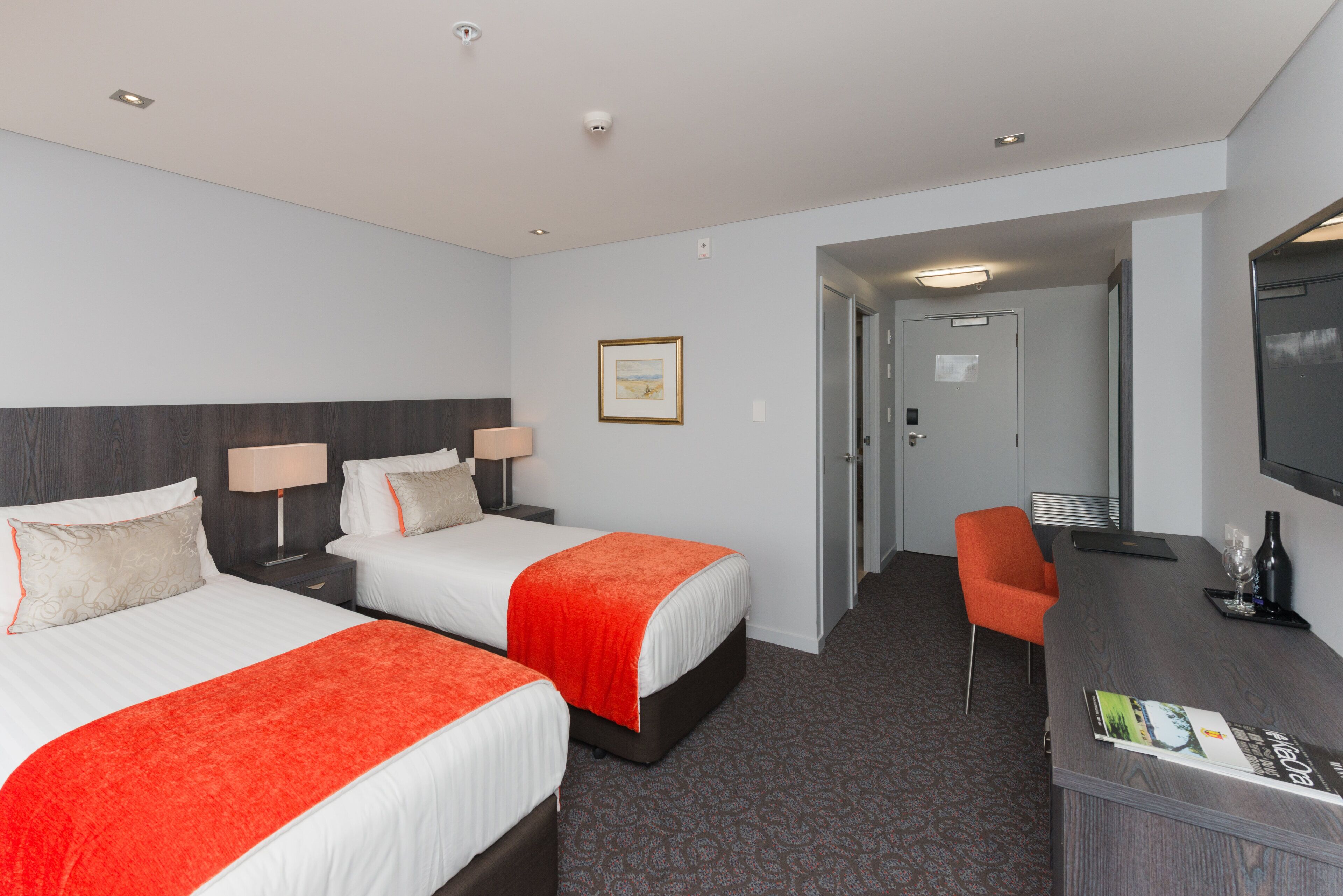 Ein Hotelzimmer mit zwei Betten, einem Schreibtisch mit Stuhl, einem Fernseher und einem gerahmten Bild an der Wand.