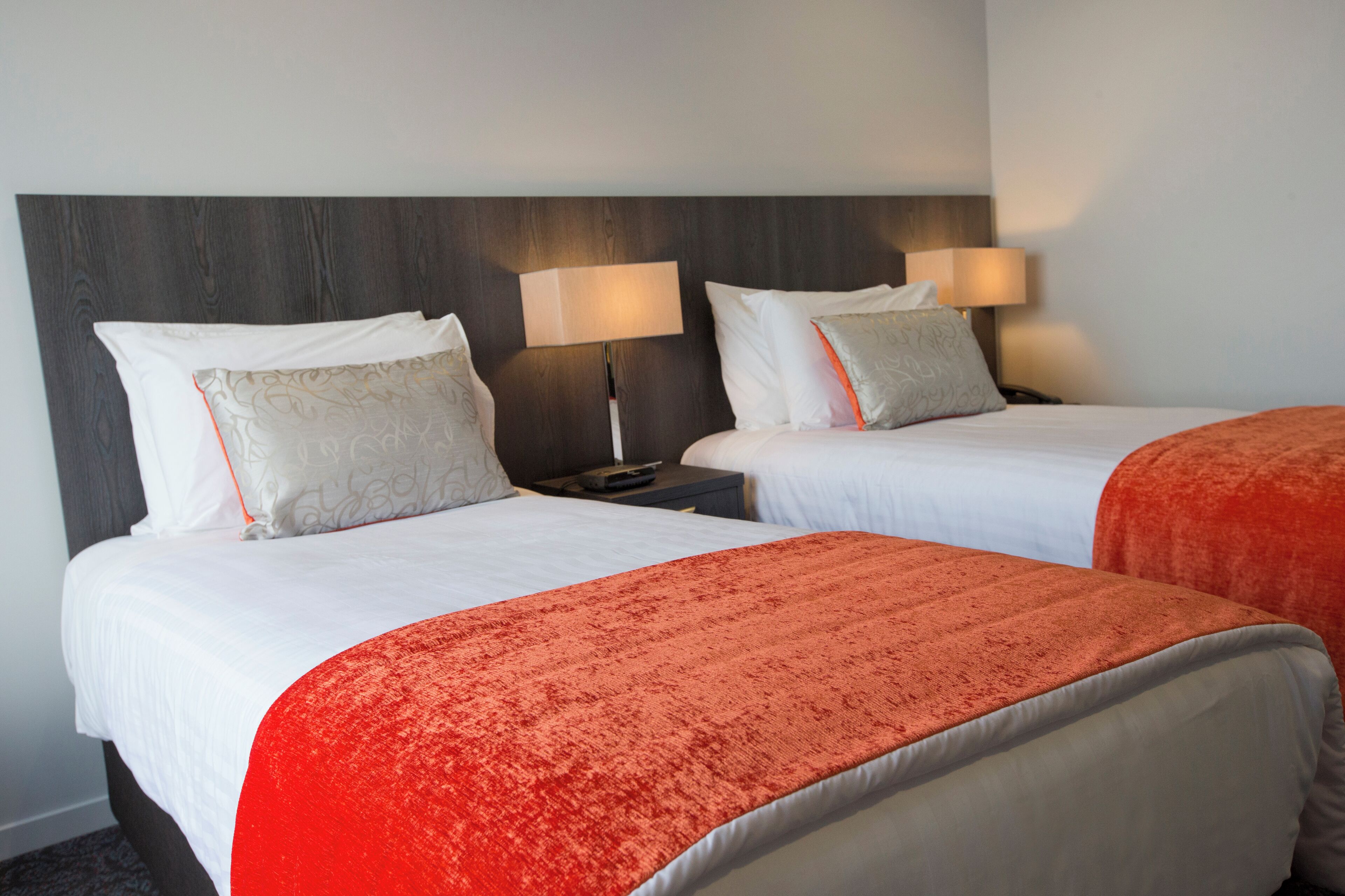 Habitación de hotel con dos camas, cada una con ropa blanca y mantas naranjas. Hay dos mesitas de noche con lámparas y un cabecero de madera oscura.