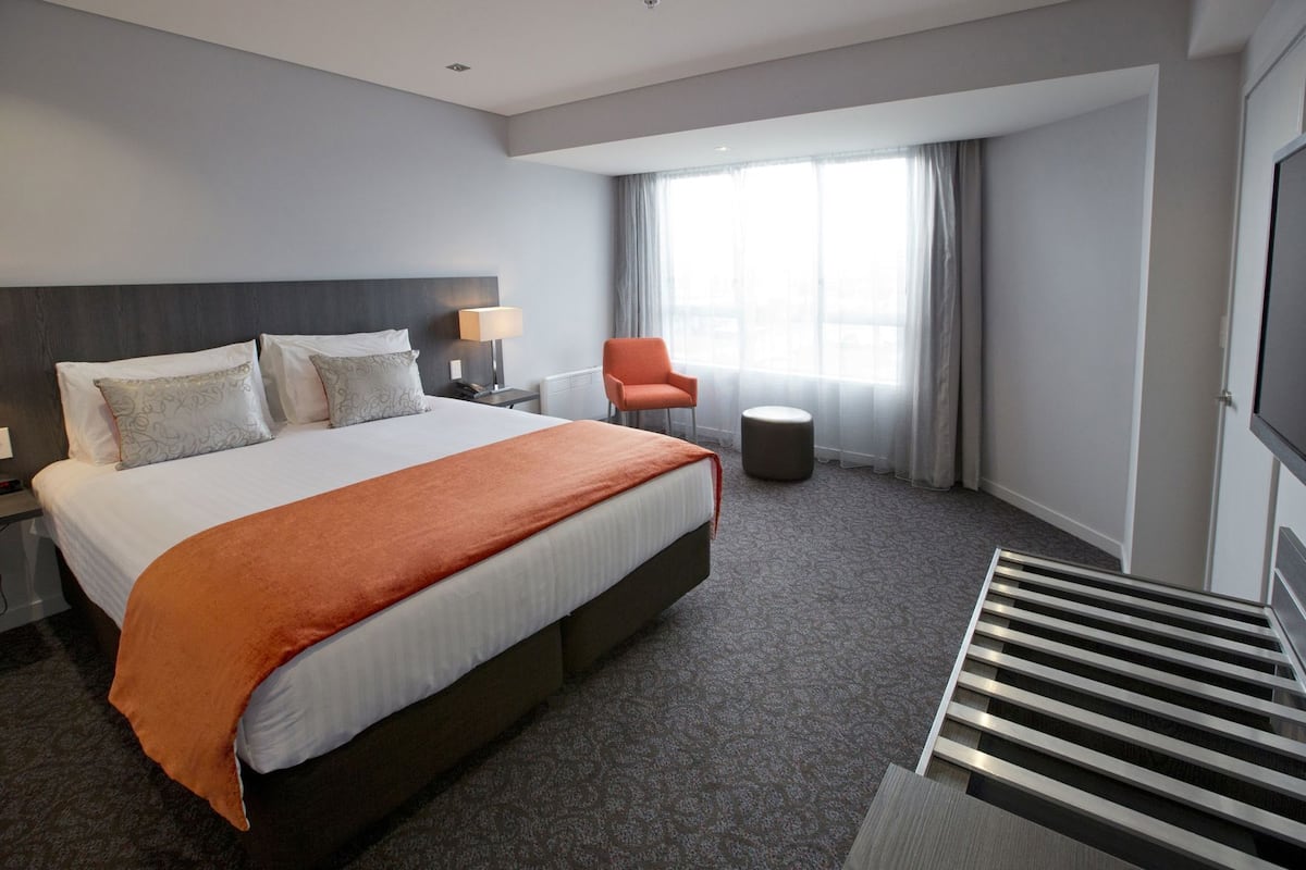 Ein Hotelzimmer mit einem großen Bett, einem Nachttisch, einem Sessel, einem kleinen Hocker und einem an der Wand befestigten Fernseher.
