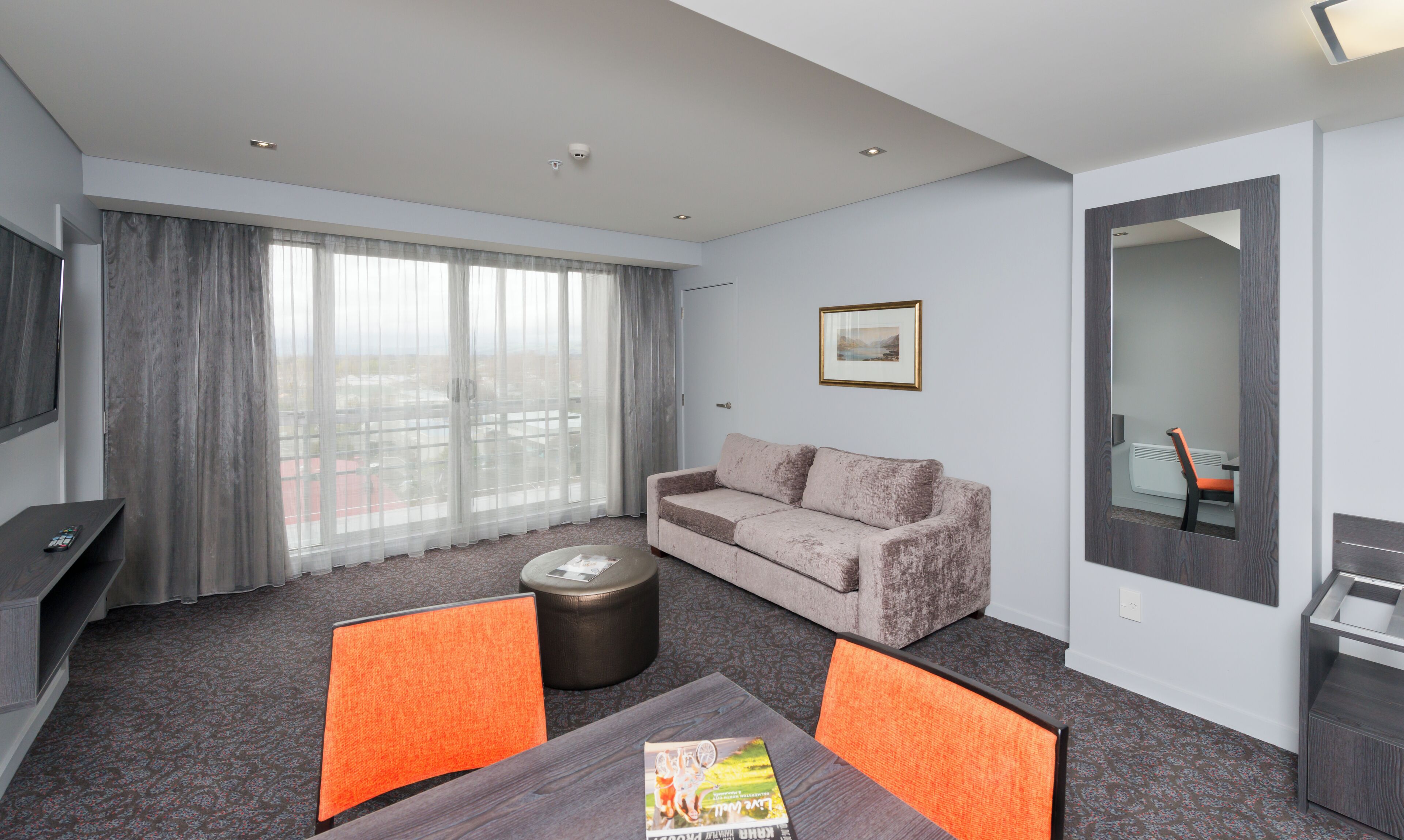 A modern hotel room with a sofa, a coffee table, a TV, and a large window with curtains.