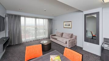 A modern hotel room with a sofa, a coffee table, a TV, and a large window with curtains.