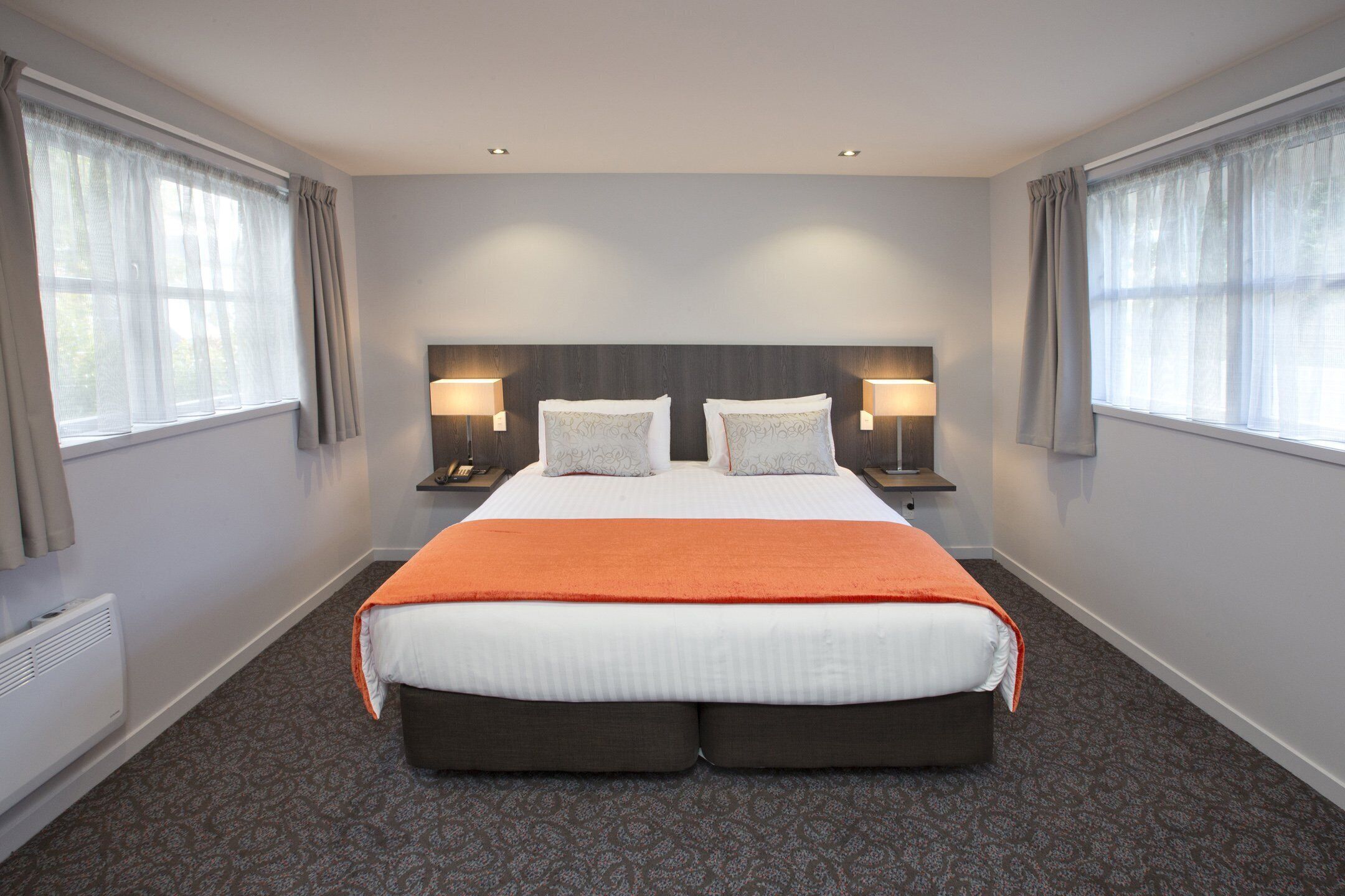 Een groot bed met een oranje deken, twee nachtkastjes met lampen, en een hoofdbord.