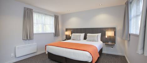 A hotel room with a large bed, two bedside tables with lamps, a window with curtains, and a radiator.