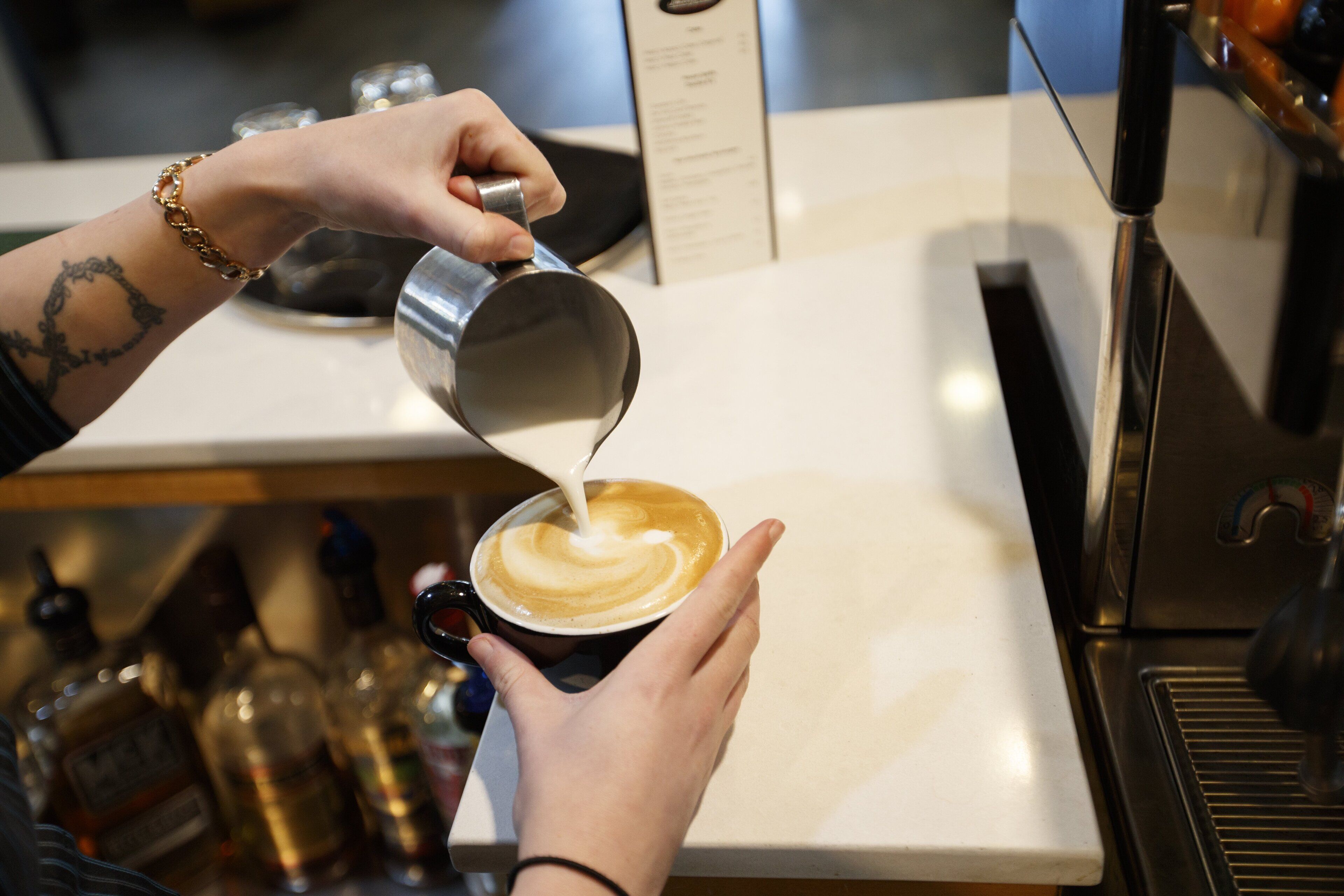 Uma pessoa despejando leite numa chávena de café, com uma máquina de café e várias garrafas ao fundo.