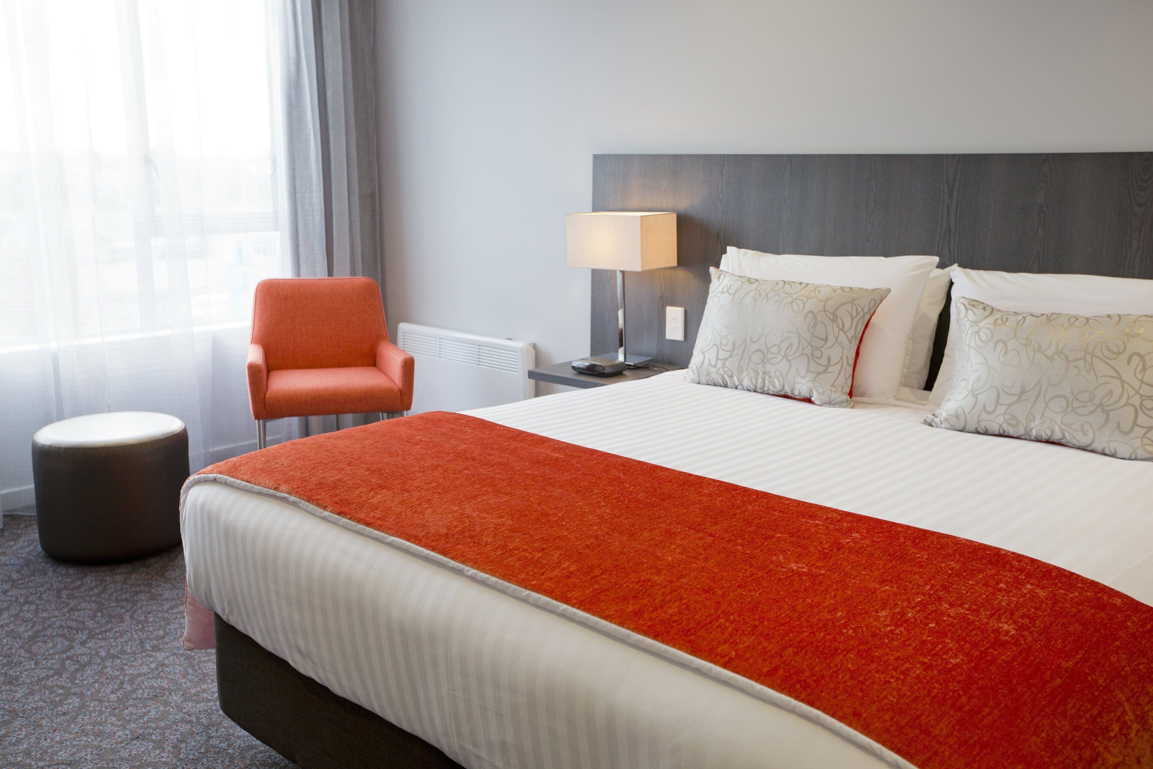 A hotel room with a bed, an orange chair, a nightstand with a lamp, and a window with curtains.