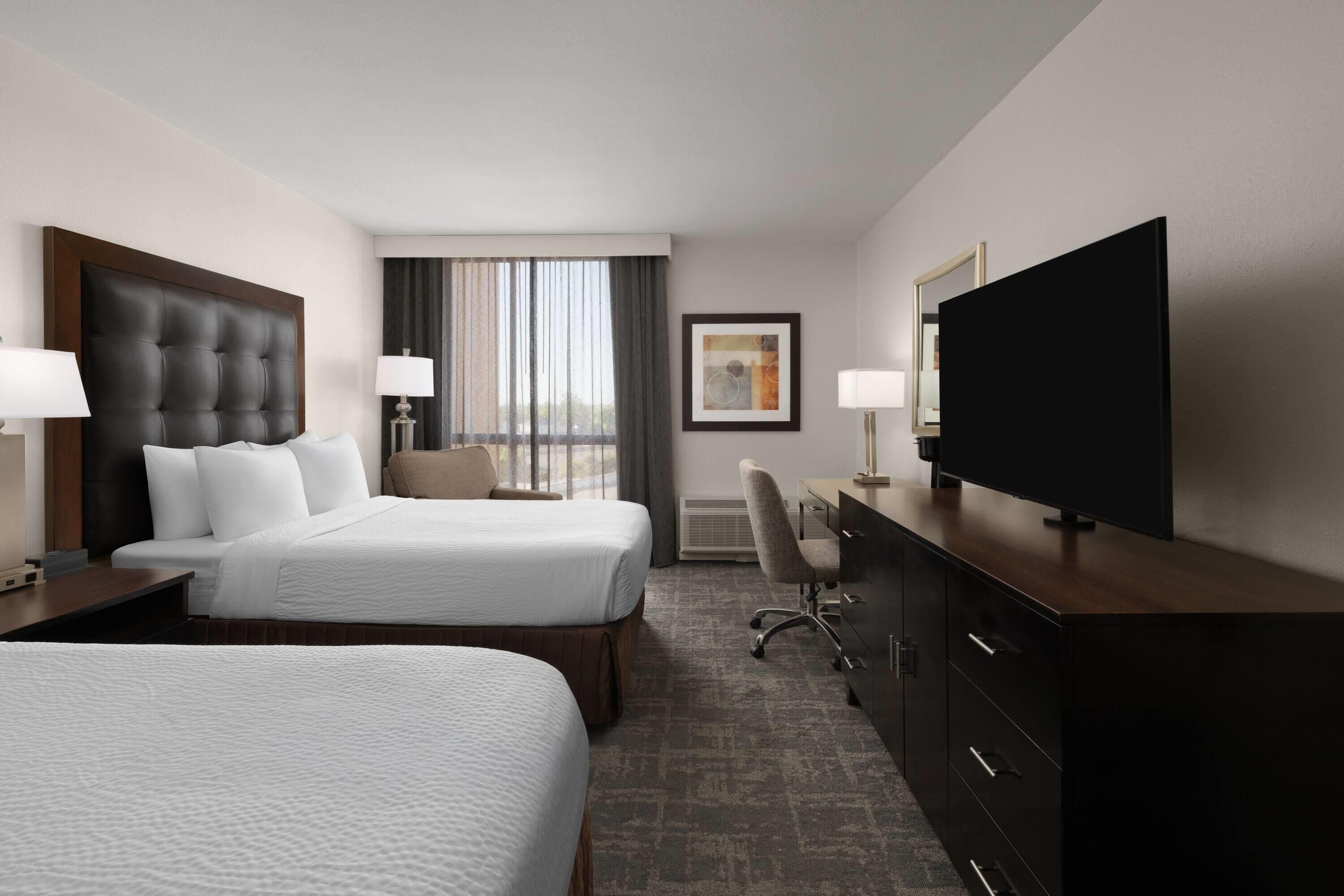 A hotel room with two beds, a desk, a television, and a window with curtains.