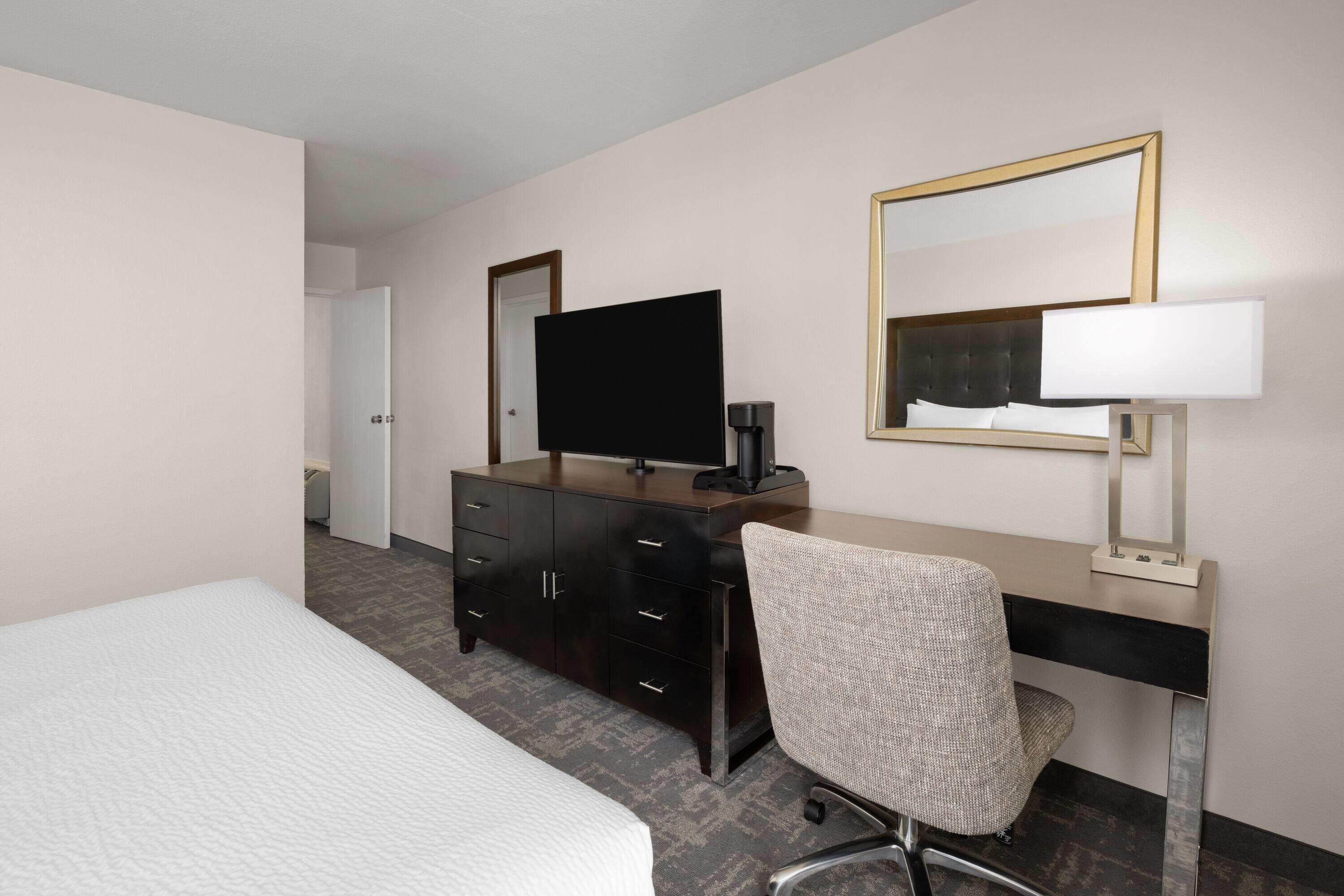 A hotel room with a bed, a desk with a chair, a television, a coffee maker, and a mirror.