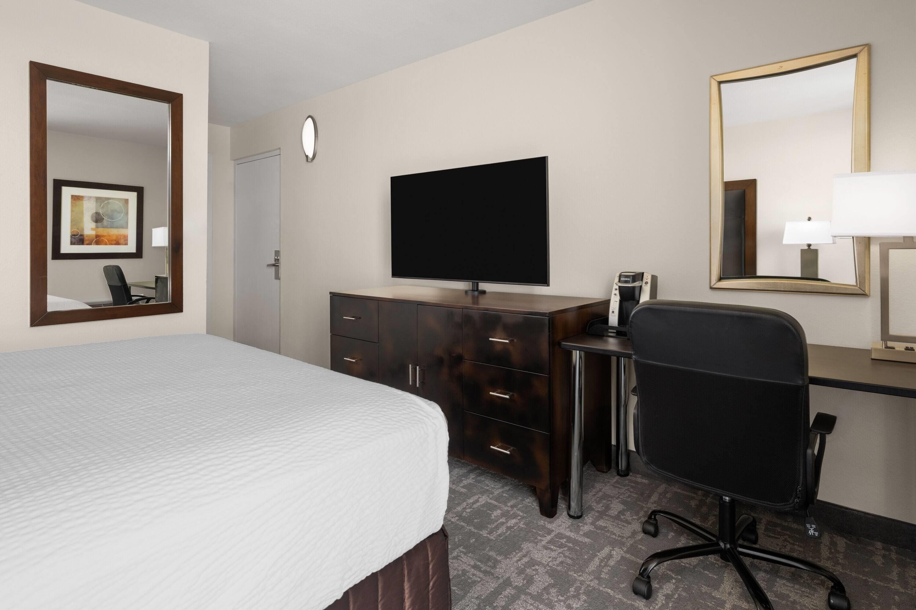 Ein Hotelzimmer mit Bett, Schreibtisch, Stuhl, Fernseher und Spiegel.