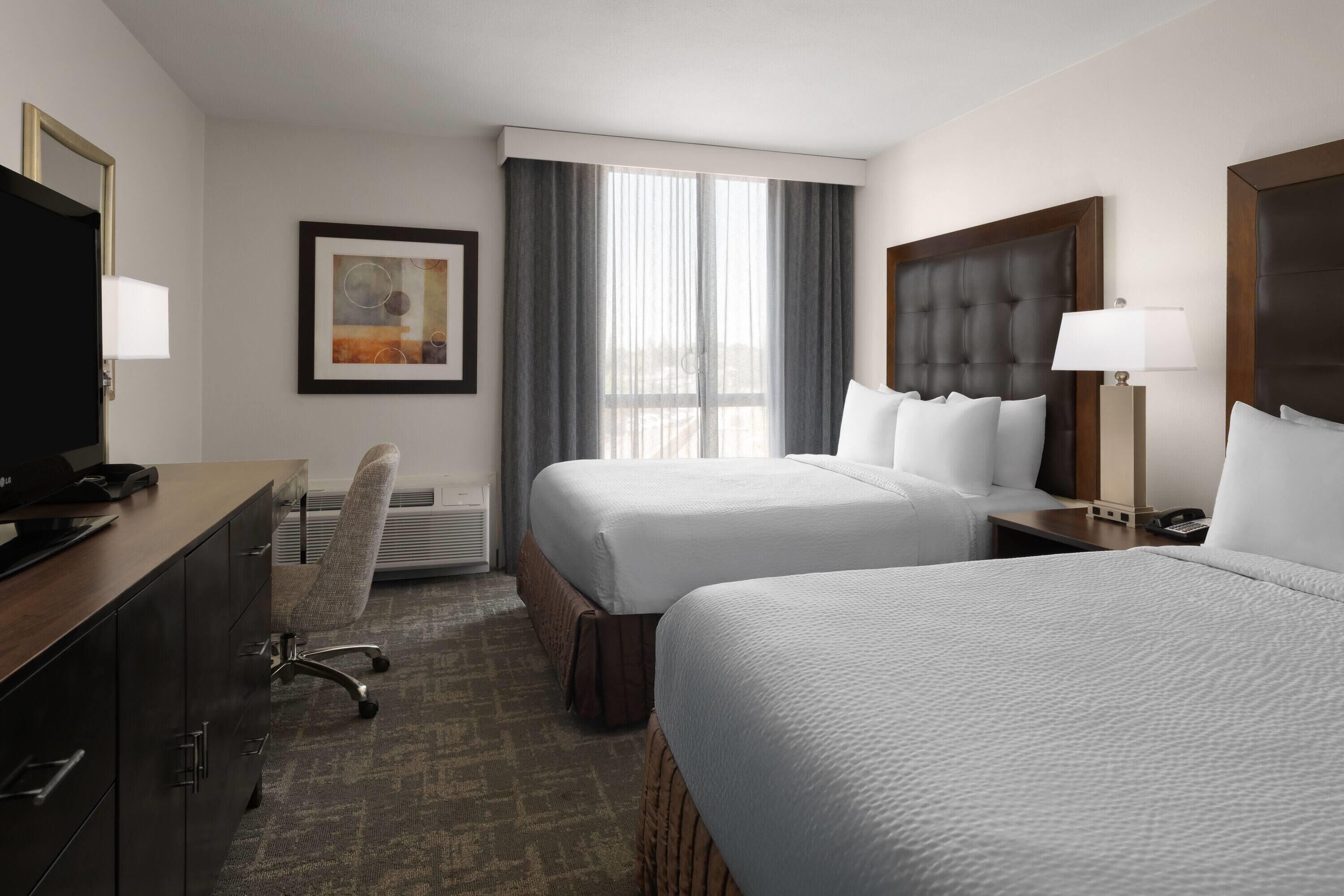 A hotel room with two beds, a desk with a TV, a chair, a framed picture, and a window with curtains.