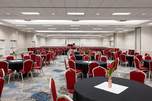 Uma ampla sala de conferências com fileiras de mesas e cadeiras, um palco com a bandeira americana e piso coberto por carpete.