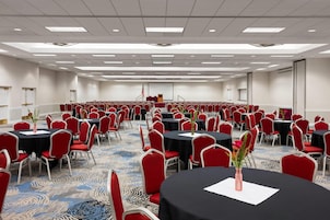 Un salón de conferencias amplio con filas de mesas y sillas, un escenario con la bandera estadounidense y suelo alfombrado.